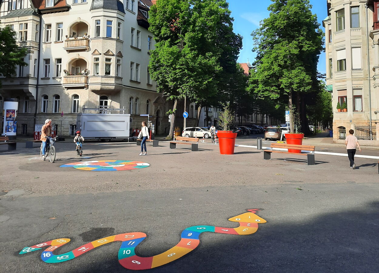 Liviaplatz in Leipzig. Im Vordergrund ist ein buntes Springspiel für Kinder auf den Asphalt markiert. Den Platz rahmen Sitzbänke, rote Pflanzkübel, Straßenbäume und Gründerzeithäuser