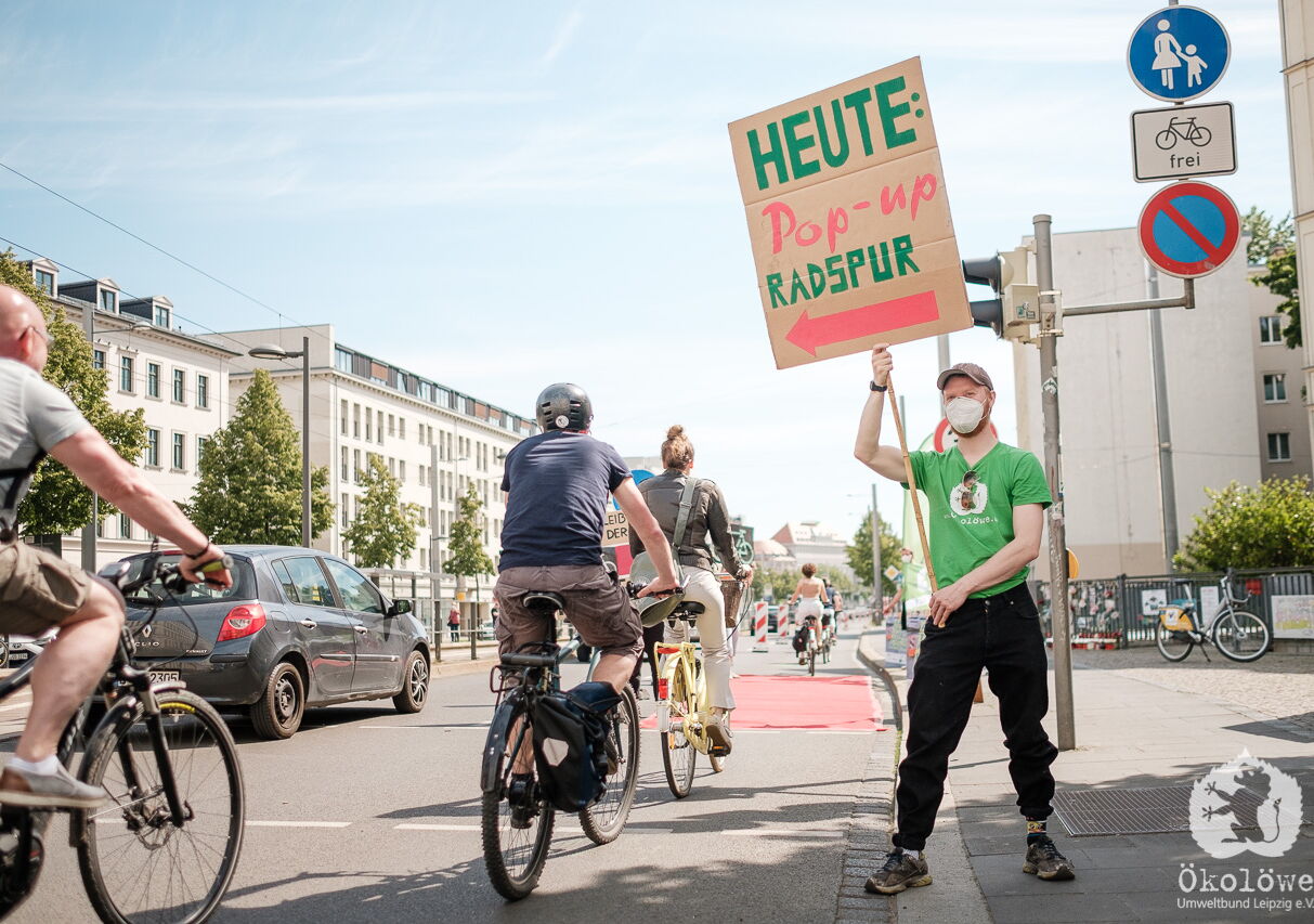 Mann im Ökolöwen T-Shirt steht neben improvisiertem Radweg und hält Schild hoch mit Aufschrift: "Heute Pop-up Radspur" Über den Radweg fahren hintereinander fünf Fahrradfahrer. Links daneben fährt ein Auto
