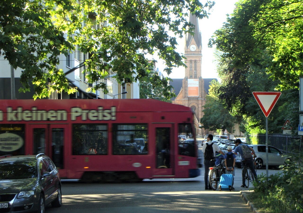 Eine Gruppe Fußgänger:innen und Radfahrer:innen steht in der Schreberstraße und möchte die Käthe-Kollwitz-Straße queren. Eine Straßenbahn fährt bedrohlich von links nach rechts.
