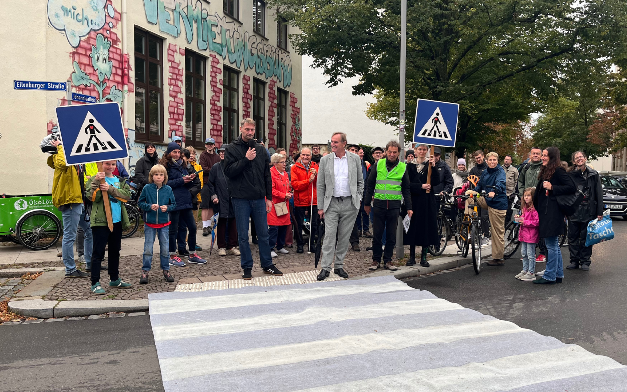 Eine Gruppe von Menschen steht vor einem selbst gebasteltem Fußgängerüberweg. Zwei Personen halten Zebrastreifen-Schilder hoch.
