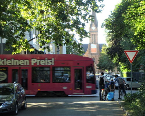 Eine Gruppe FußgängerInnen und RadfahrerInnen steht in der Schreberstraße und möchte die Käthe-Kollwitz-Straße queren. Eine Straßenbahn fährt bedrohlich von links nach rechts.