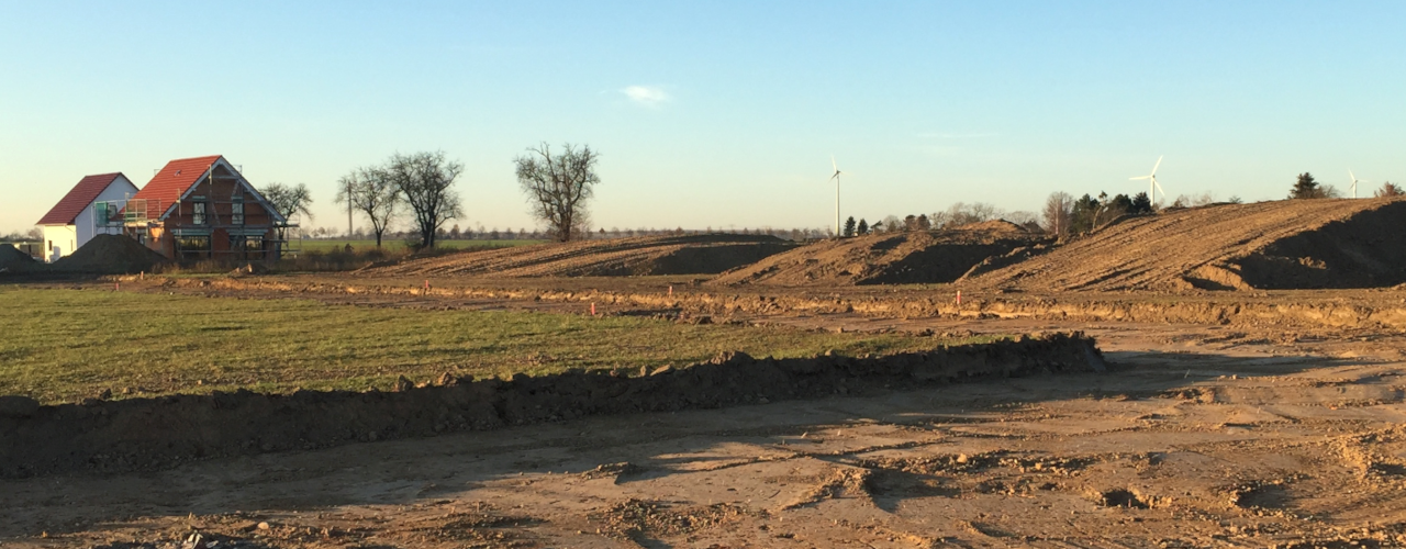 Auf dem Bild ist im Vordergrund eine fein säuberlich ausgehobene Baugrube. Daneben sieht man noch den Acker, der hier bebaut werden soll. Rechts sind Hügel mit Erde. Links zwei Einfamilienhäuser mit Baugerüst.