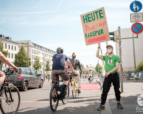 Mann im Ökolöwen T-Shirt steht neben improvisiertem Radweg und hält Schild hoch mit Aufschrift: "Heute Pop-up Radspur" Über den Radweg fahren hintereinander fünf Fahrradfahrer. Links daneben fährt ein Auto