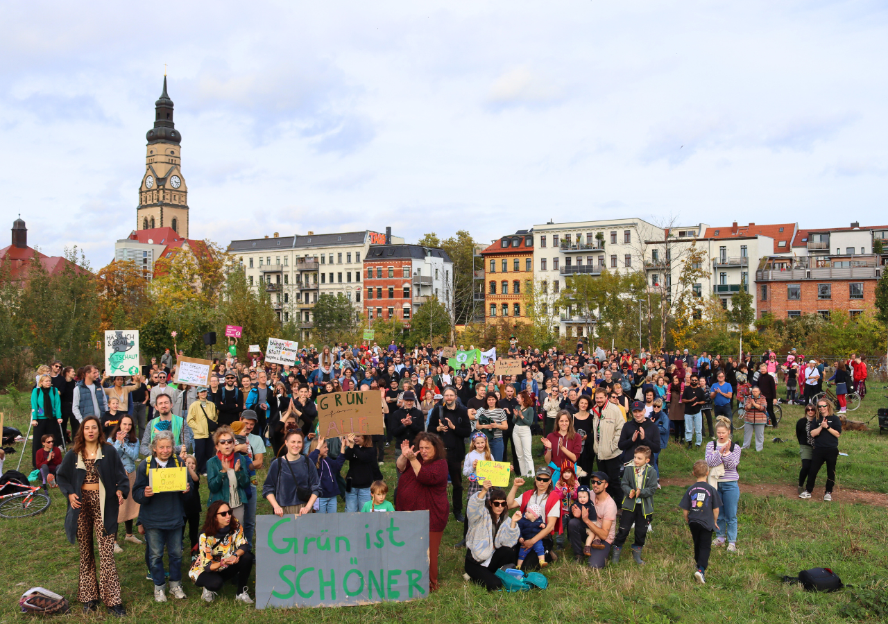 Eine große Gruppe Demonstranten steht auf dem Jahrtausendfeld in Leipzig Plagwitz und hält Schilder hoch. Im Hintergrund eine Kirche und Wohnhäuser