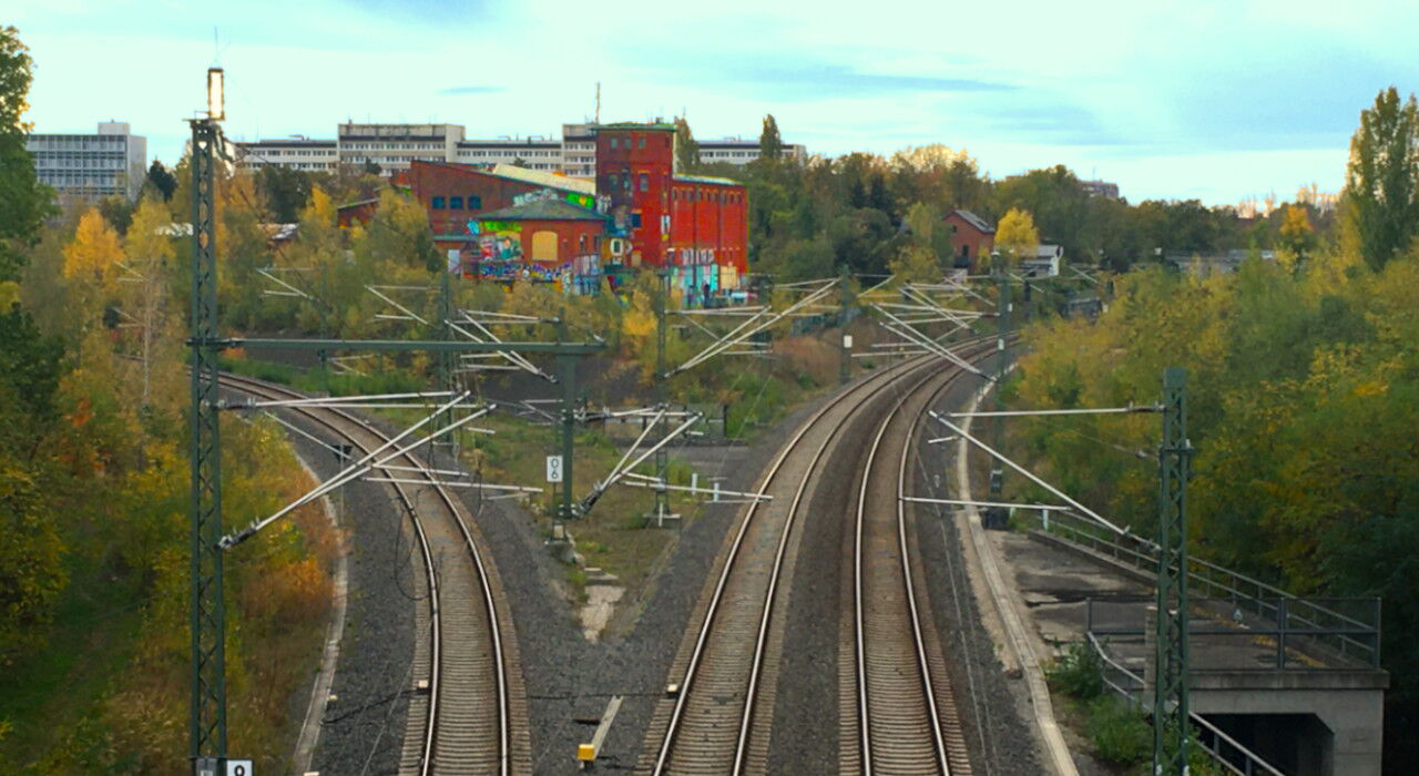 Bahngleise gabeln sich. In der Mitte erhebt sich ein großer Backsteinbau mit viel Graffiti an der Fassade. Die Bahndämme im Umfeld sind mit viel Grün zugewachsen
