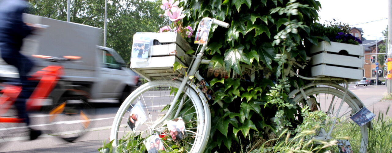 Ghostbike, weiß angemaltes Fahrrad mit handgeschriebenen Schildern in Andenken an eine getötete Radfahrerin steht neben einer Straße mit Verkehr.