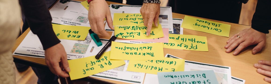 Handgeschriebene Zettel auf einem Tisch, Menschen stehen drumherum und zeigen auf die Zettel