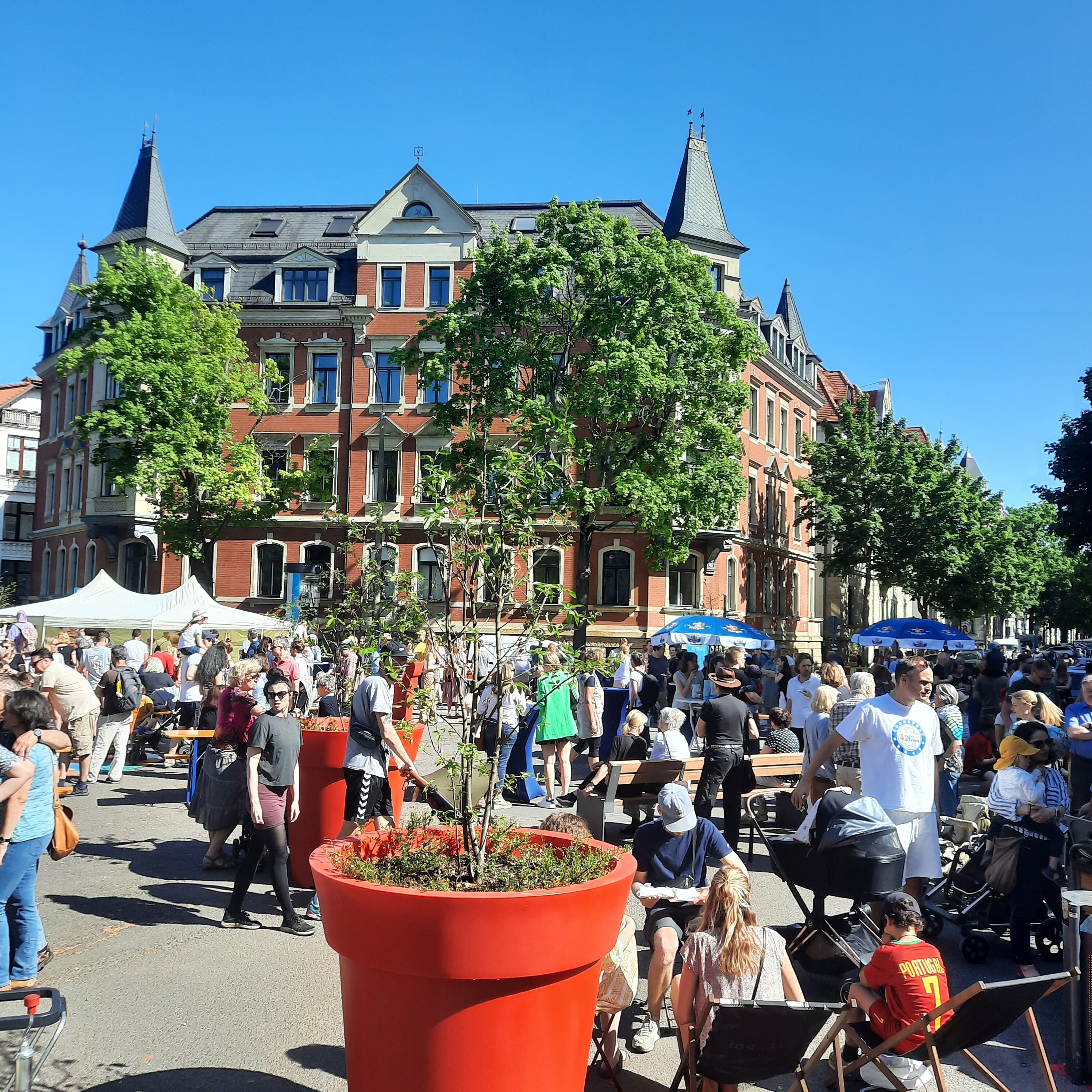 Im Vordergrund ein roter Blumenkübel. Dahinter ein Platz voller Menschen mit Liegestühlen, Sonnenschirmen. Im Hintergrund große Straßenbäume und ein prächtiges Gründerzeithaus.