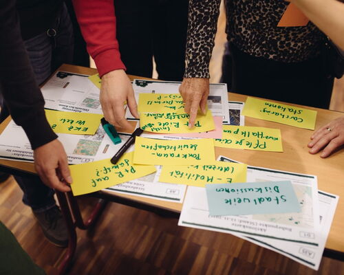 Handgeschriebene Zettel auf einem Tisch, Menschen stehen drumherum und zeigen auf die Zettel