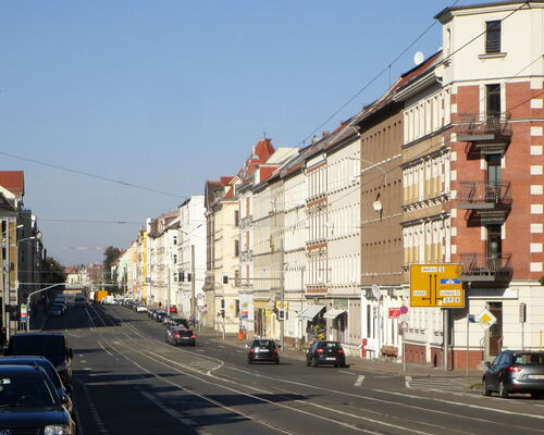 mehrspurige Georg-Schumann-Straße mit autos aber ohne Radwege und Straßenbäume. Die Sonne scheint an die Gründerzeithäuser rechts und links im Bild.