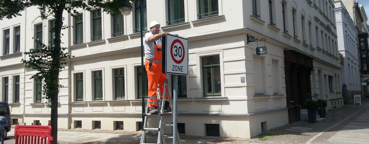 Ein man in orangener Latzhose steht auf einer Leiter und schraubt ein Tempo-30-Zonen-Schild an. Im Hintergrund ein Straßenbaum und eine Gründerzeithaus
