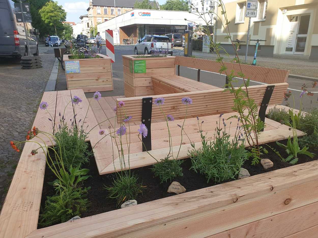 Im Vordergrund eine frisch aufgebaute Sitzgelegenheit aus Holz. Rings herum sind in dem Holzkasten Blumen angepflanzt. Im Hintergrund ist eine Straße mit Autos und der Konsum Supermarkt im Bachviertel zu sehen