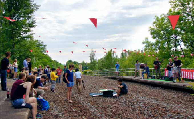 In der Mitte verläuft ein Schottergleisbett ohne Gleise. Im Schotterbett und links und recht an den Bahnsteigkanten sitzen Menschen. Darüber sind Wimpelketten gespannt.
