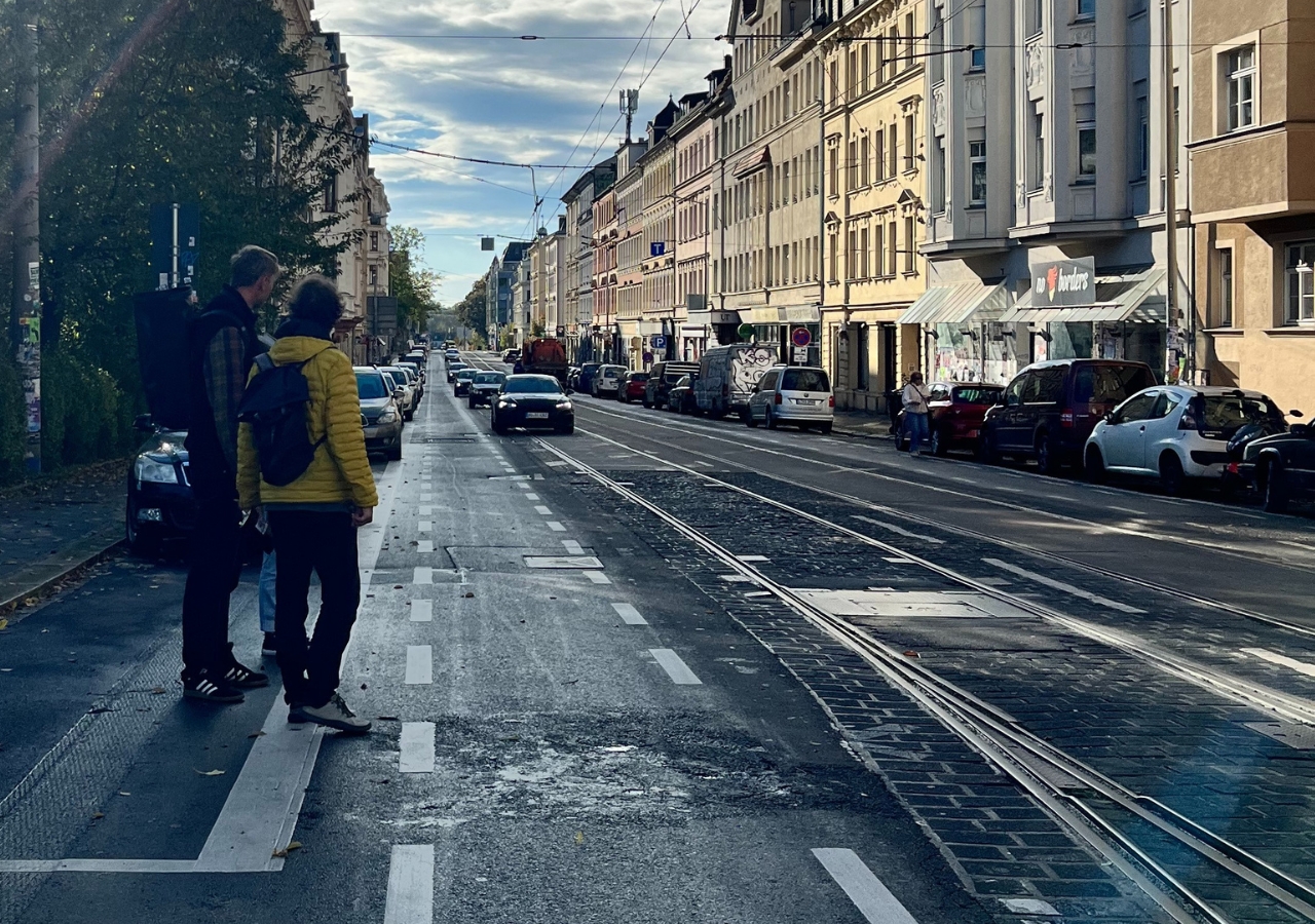 Zwei Personen mit Rucksäcken stehen an einer Straße mit Straßenbahnschienen, umgeben von mehrstöckigen Gebäuden und parkenden Autos. Die Personen versuchen die vielbefahrene Straße zu überwueren.
