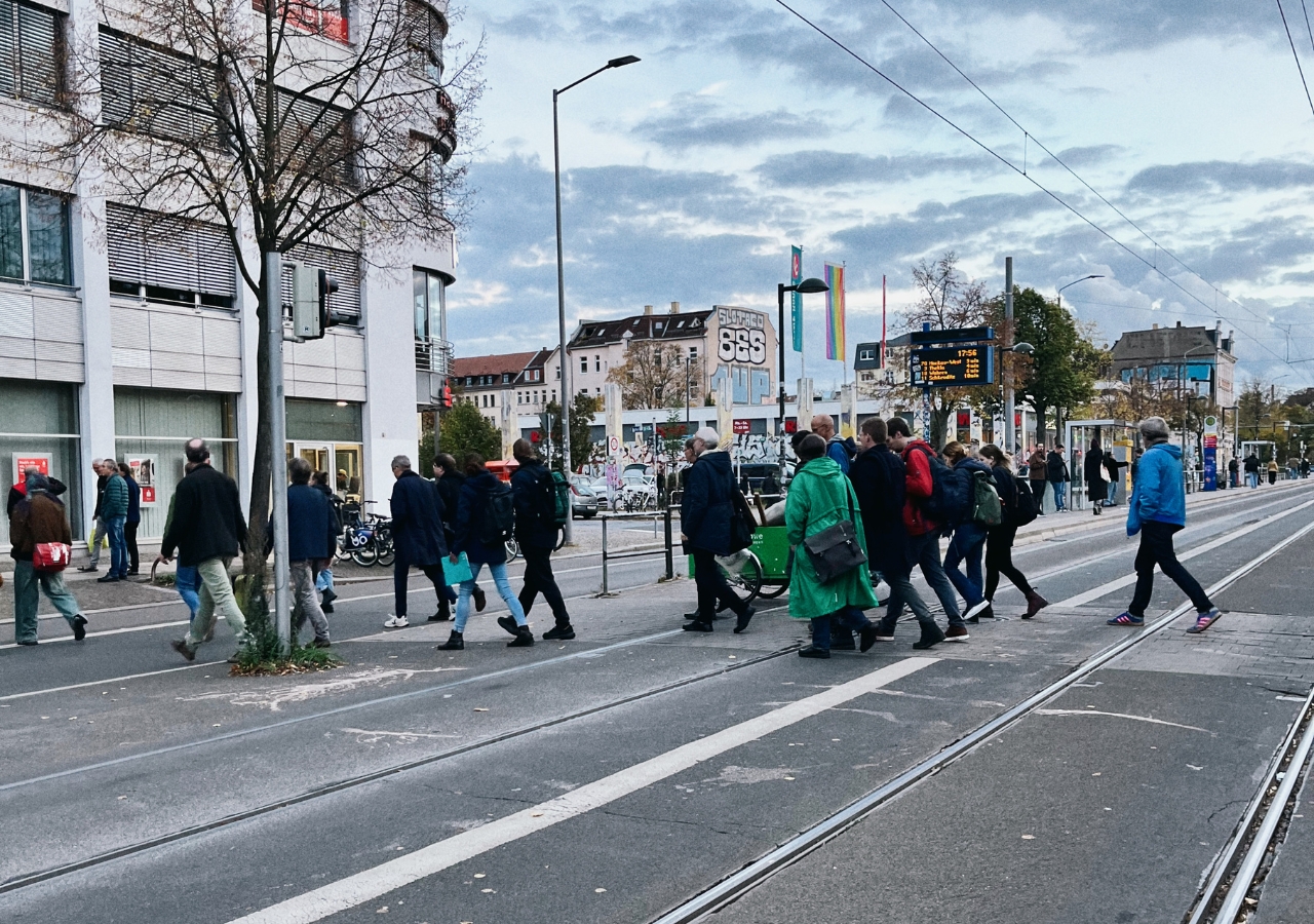 Menschen überqueren eine vielbefahrene Straße mit Straßenbahnschienen in einer städtischen Umgebung bei bewölktem Himmel.