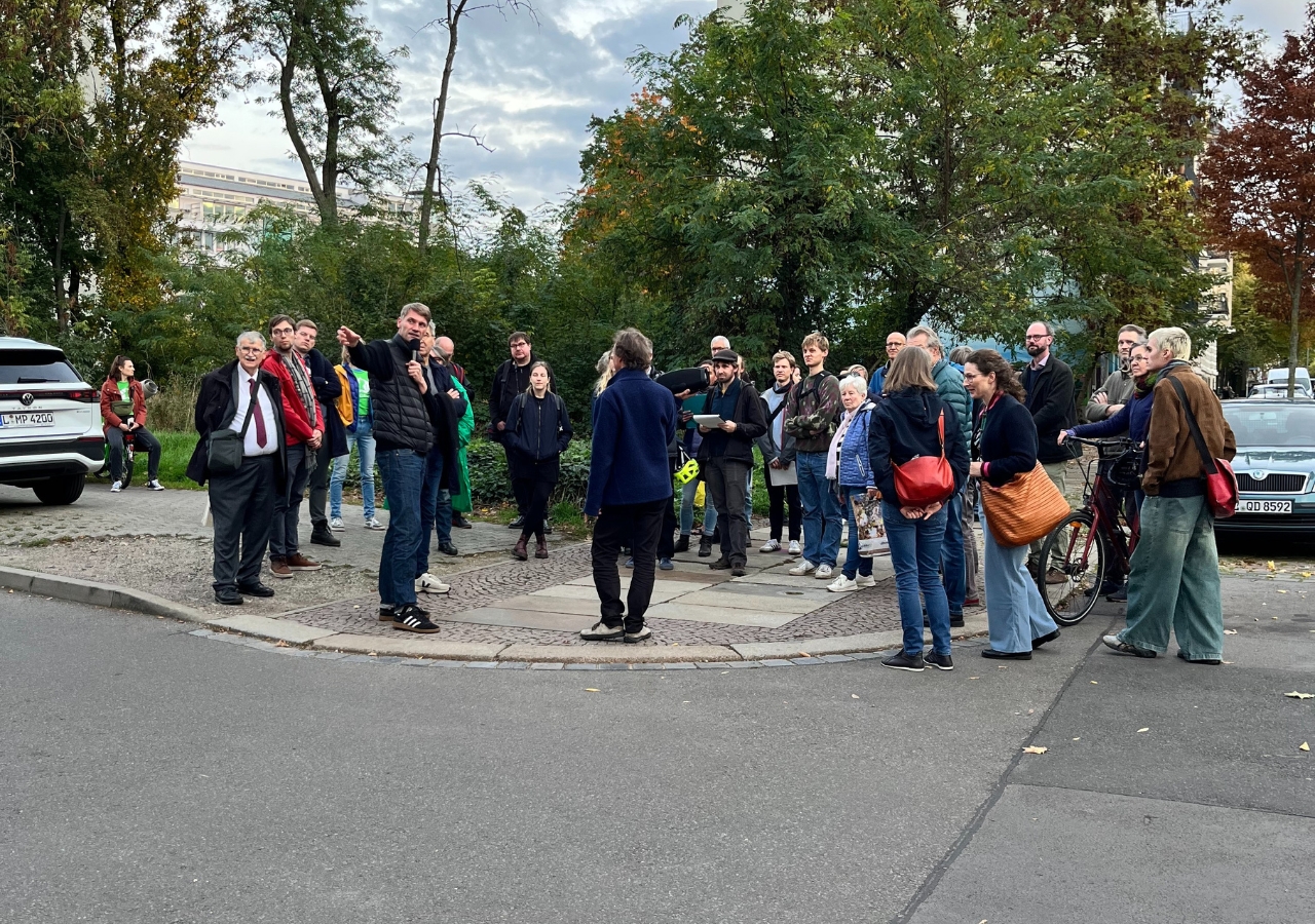 Gruppe von Menschen steht auf einem gepflasterten Kreisplatz an einer Kreuzung, umgeben von Bäumen und parkenden Autos.