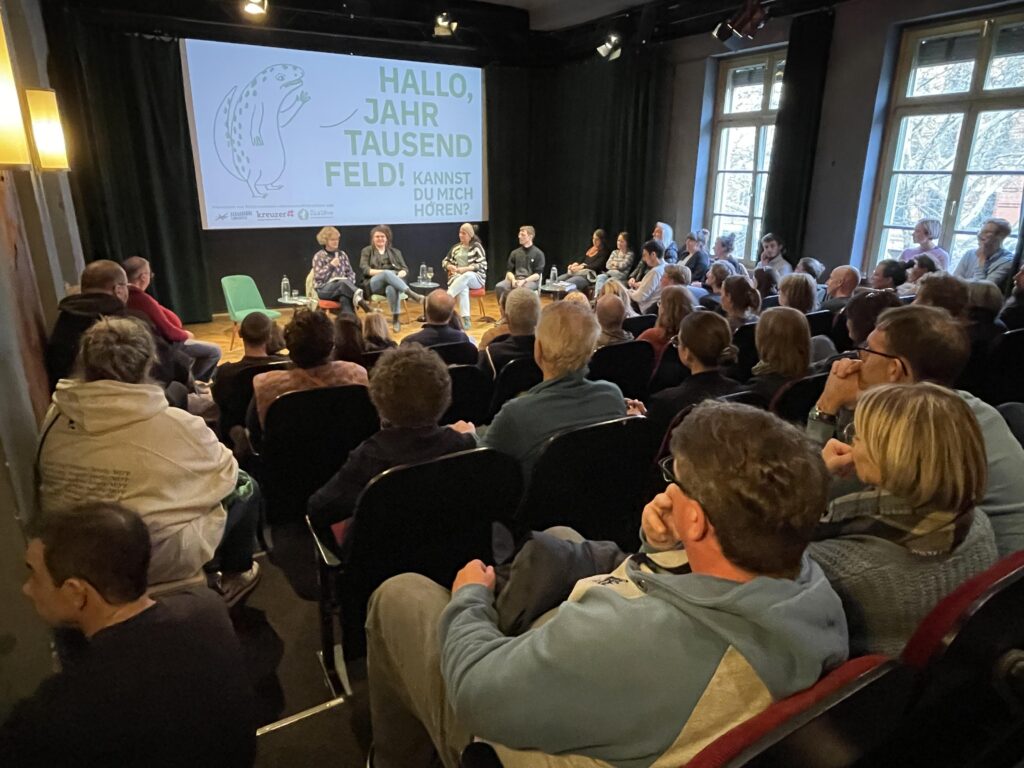 Auf einer Bühne in der Schaubühne Lindenfels sitzen 4 Menschen und diskutieren. In den voll besetzten Stuhlreihen davor hören die Menschen zu. An die Leinwand ist ein Bild geworfen auf dem steht "Hallo Jahrtausendfeld! Kannst du mich hören?"
