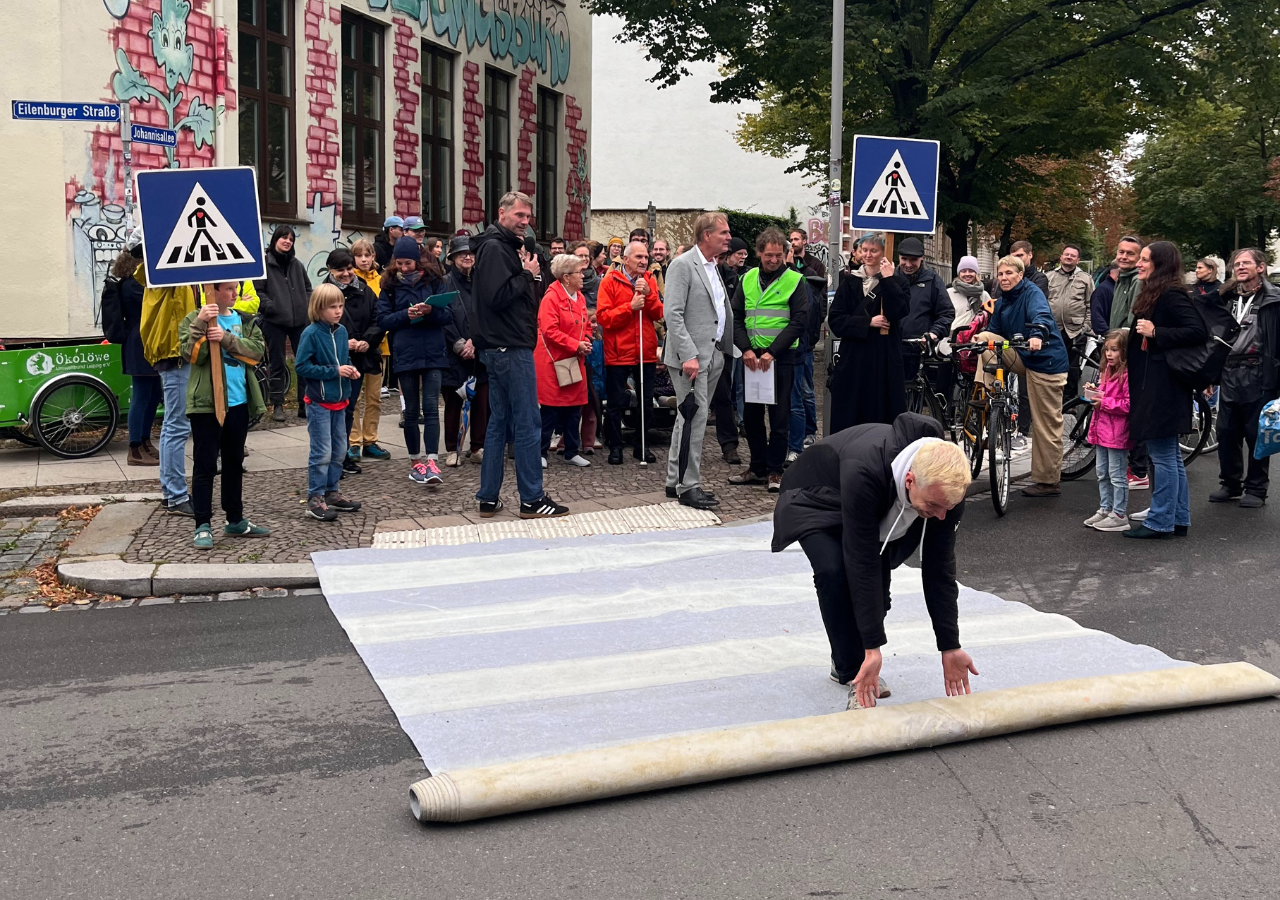 Wir Ökolöwen rollen in der Eilenburger Straße schon mal den Zebrastreifen aus. Drumherum steht eine Gruppe Menschen.