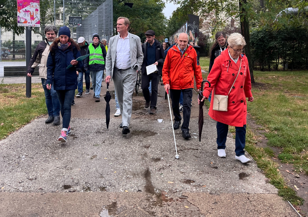 Eine Gruppe Menschen läuft auf einem Gehweg im Lene-Voigt-Park mit Schlaglöchern. Eine älteres Pärchen mit roten Jacken auf der rechten Seite. Der Mann hat einen Blinden-Stock in der Hand