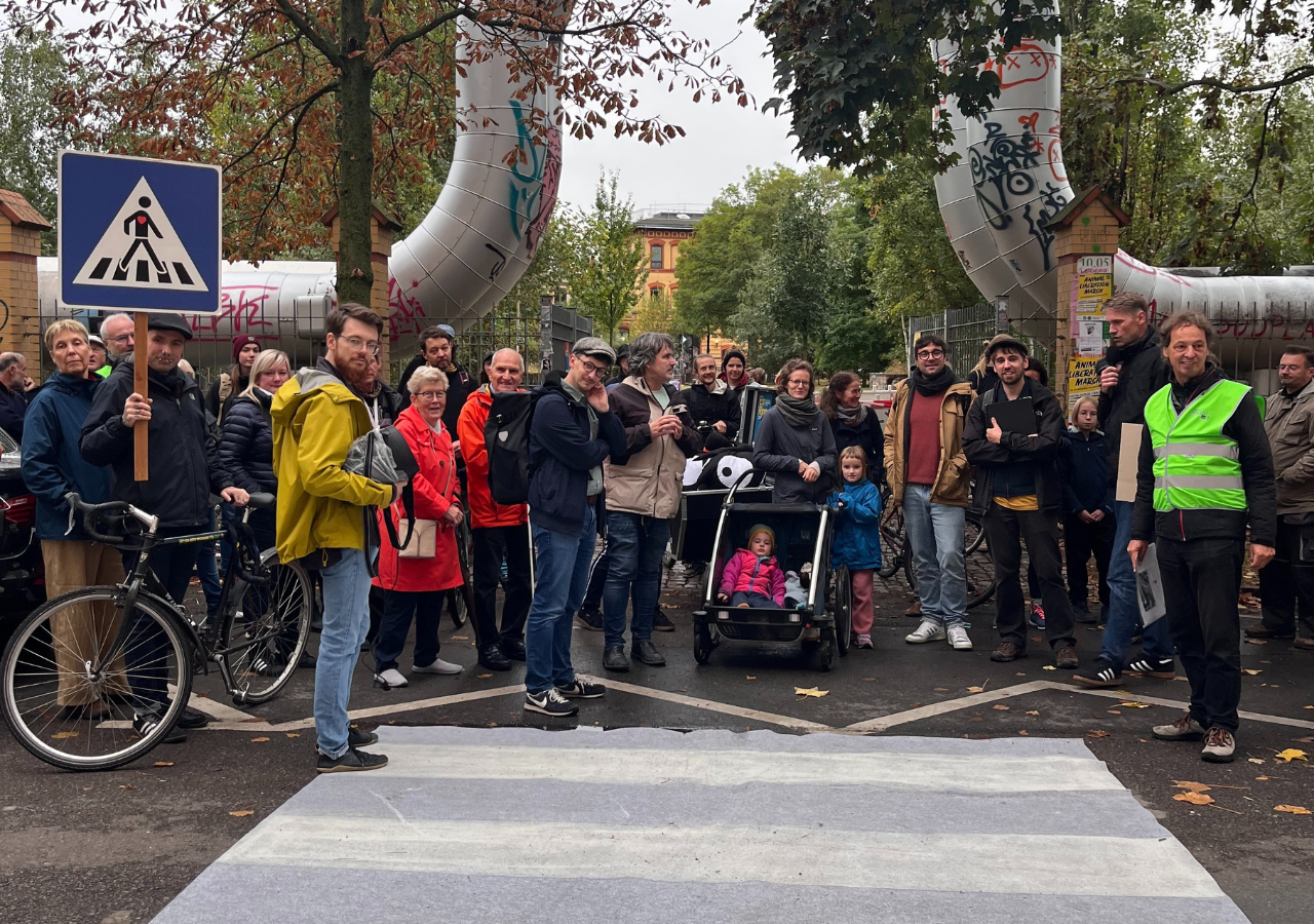 Eine Gruppe von Menschen steht vor einem selbst gebasteltem Fußgängerüberweg. Zwei Personen halten Zebrastreifen-Schilder hoch. Im Hintergrund eine Fernwärmeleitung