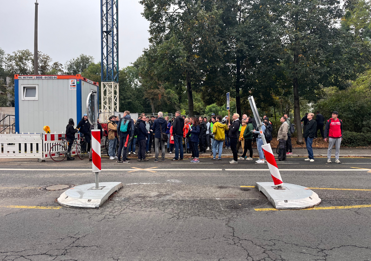 Eine Gruppe von Menschen stehen vor einer temporären Querungshilfe in der Straßenmitte. Links im Hintergrund eine Baustelle. Rechts ein Park.