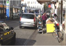 Fußgänger auf Gehweg zwischen parkenden Autos und Aussteller eines Ladens