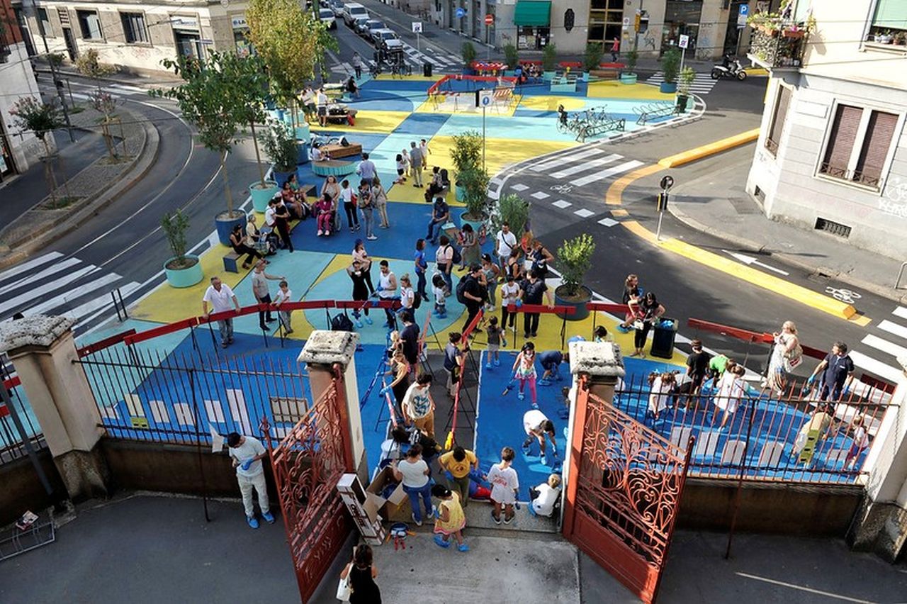 Straßenkreuzung in Mailand die mit blauer Farbe und einfachen Einbauten zu Platz umgestaltet wurde. Darauf spielen viele Kinder. Zum Platz führen Zebrastreifen