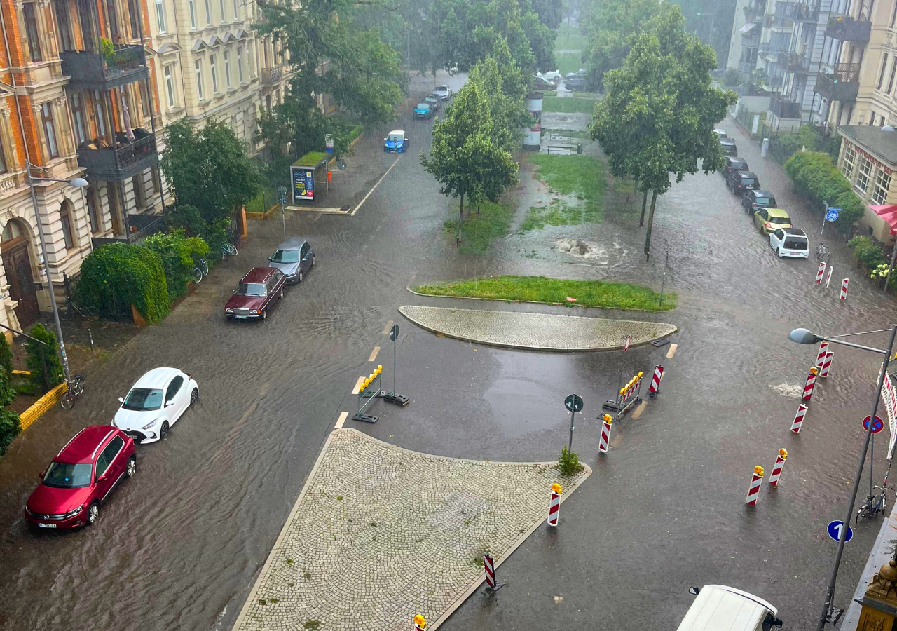 Überflutete Kreuzung mit mehreren geparkten Autos und Bäumen, Wasser bedeckt die Straße und Gehwege.