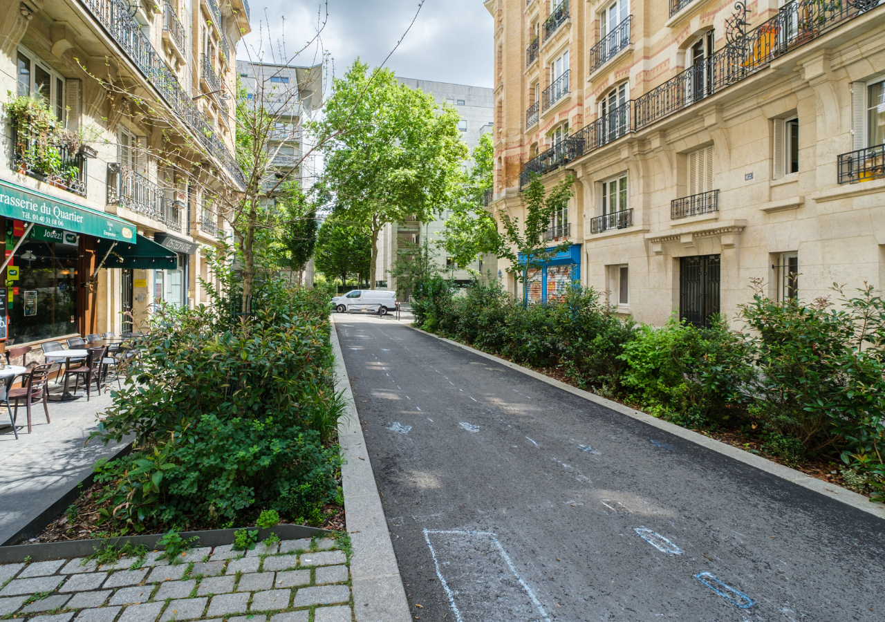 Schmale Straße in Paris mit Bäumen und Sträuchern in Pflanzbeeten entlang des Gehwegs, umgeben von mehrstöckigen Wohngebäuden.