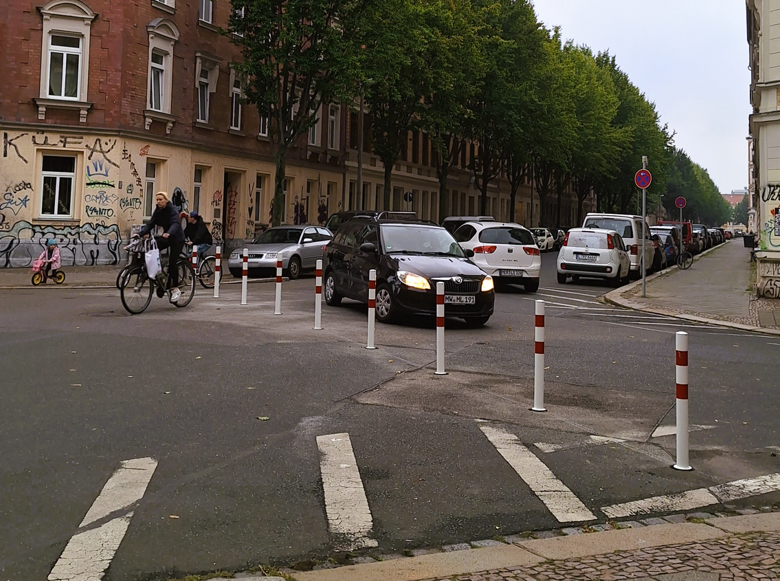 Eine Poller-Reihe geht einmal quer über die Kreuzung. Fahrradfahrer fahren da durch. Ein Auto muss an der Sperre abbiegen. Im Hintergrund eine Nebenstraße mit vielen parkenden Autos, Straßenbäumen und Gründerzeithäusern