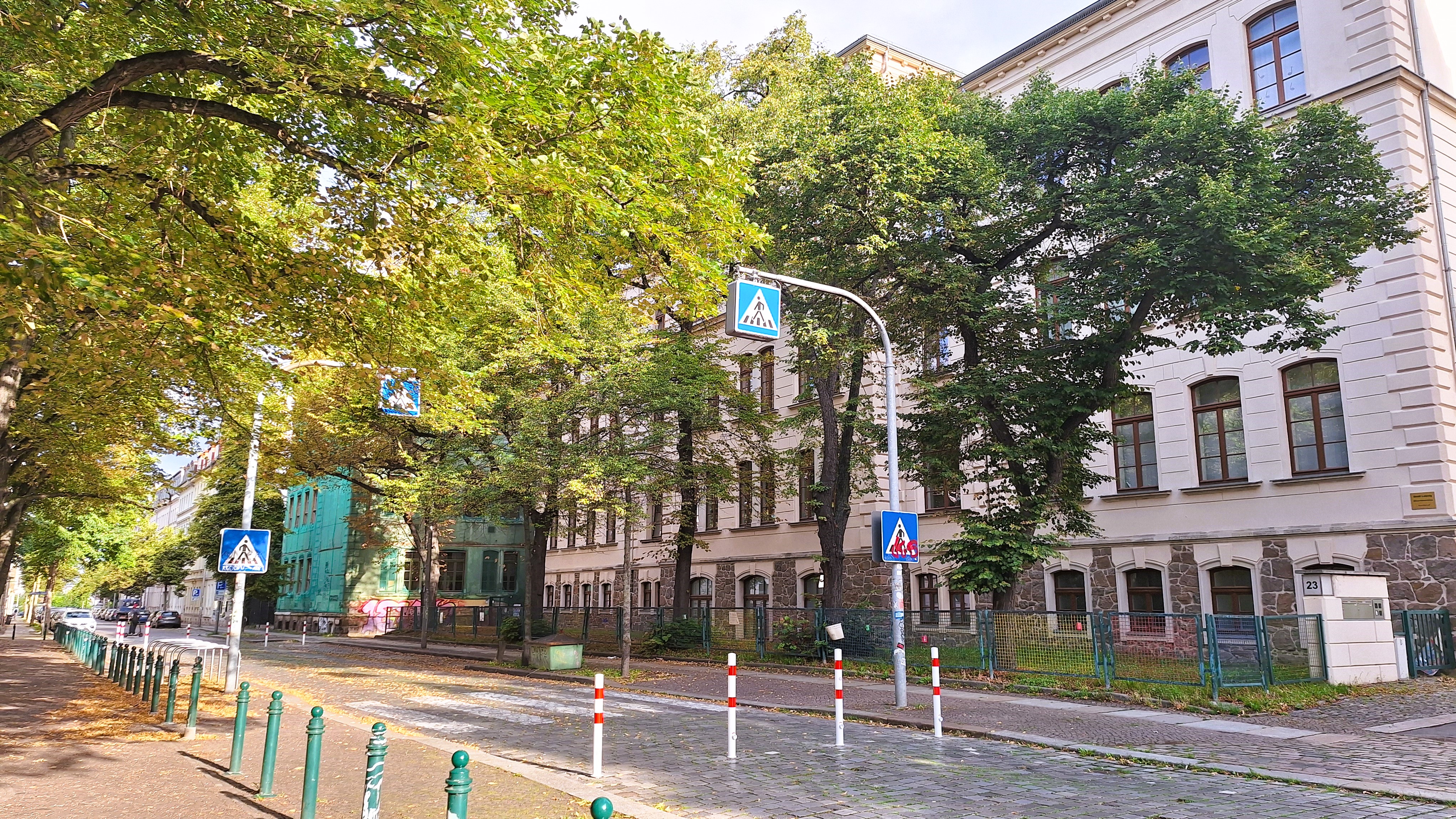 gründerzeitliches Schulgebäude mit verkehrsberuhigter Schulstraße davor. Autofrei durch Poller und Zebrastreifen. Der Belag ist Kopfsteinpflaster. Links und rechts stehen große Straßenbäume