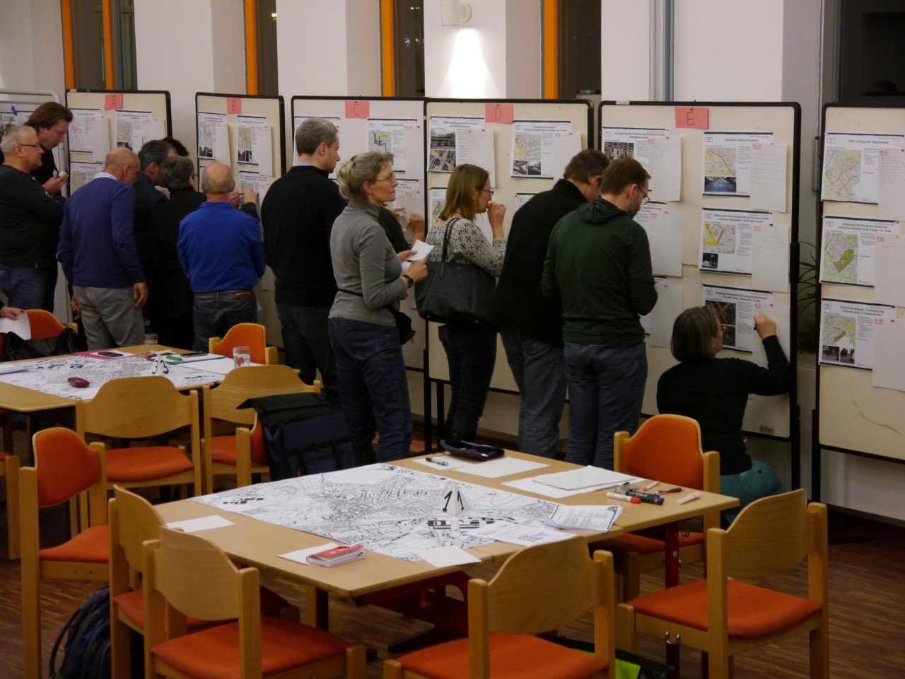 Tische mit Kartenmaterial und Menschen an Pinnwänden mit Maßnahmenblättern
