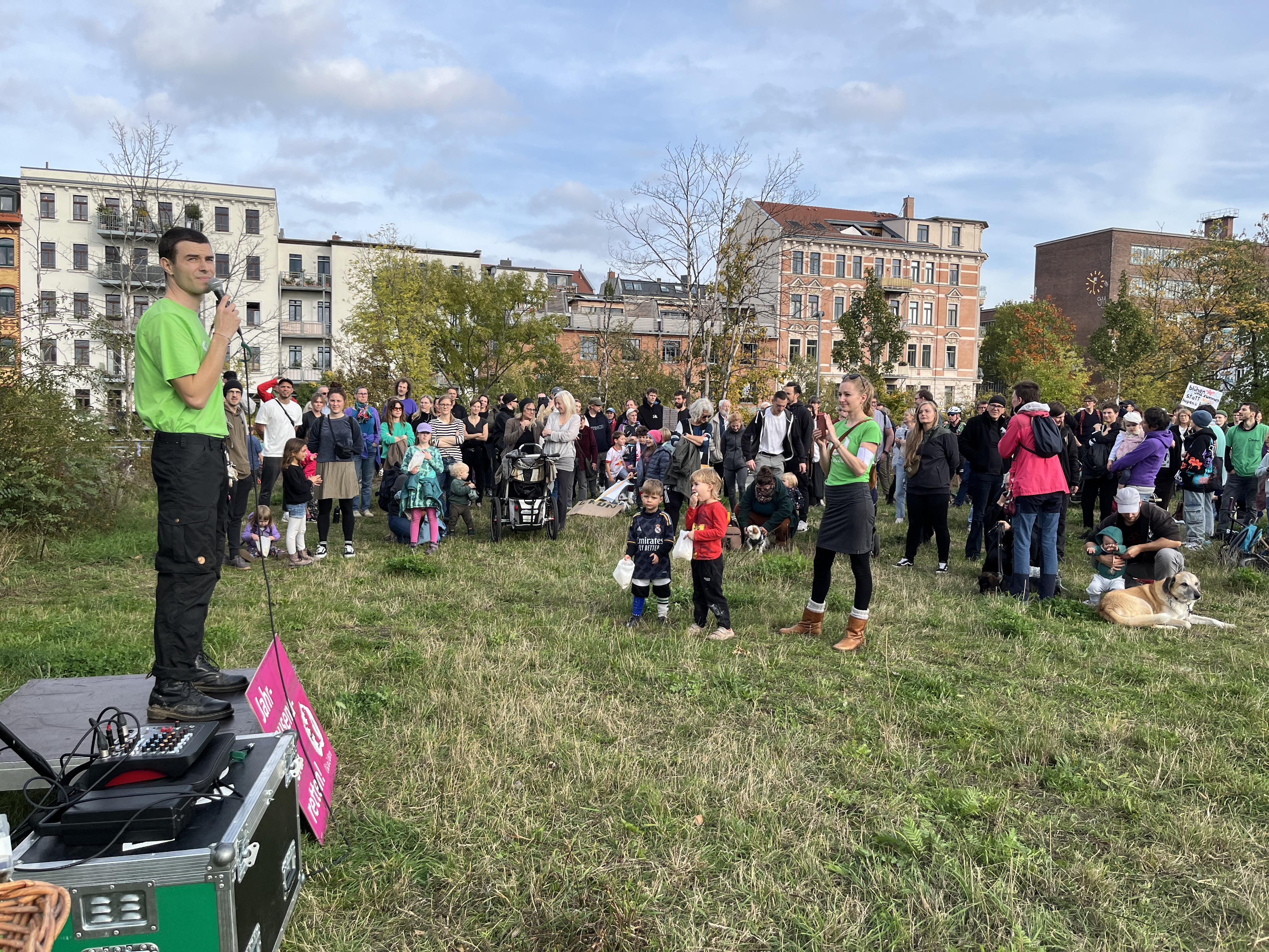 Demo auf dem Jahrtausendfeld in Leipzig, ein Mann steht auf einem Podest mit Mikrofon. Davor steht eine große Gruppe Anwohner mit Kindern auf der Wiese